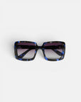 Die quadratische Sonnenbrille „ISCHIA“ von Okkia verfügt über eine schwarz-blau marmorierte Oversize-Fassung und dunkle, polarisierte Gläser mit Farbverlauf. Die Sonnenbrille wird mit der Vorderseite nach oben auf einem schlichten weißen Hintergrund präsentiert, wobei beide Bügel sichtbar sind und leicht nach außen gebogen sind.