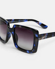 Eine Nahaufnahme der ISCHIA-Sonnenbrille zeigt das übergroße, eckige Brillengestell mit einem glänzenden, schwarz-blau marmorierten Rahmen, grauen, polarisierten Verlaufsglasen und dem in Weiß auf den Bügel aufgedruckten Markennamen „Okkia“. Das Bild präsentiert diese stilvolle Brille sowohl von der Seite als auch von vorne vor einem weißen Hintergrund.