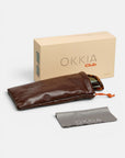 Auf einer weißen Fläche liegen eine beigefarbene Okkia Club-Box, ein braunes, gepolstertes Sonnenbrillenetui mit orangefarbenem Kordelzug, ein graues Okkia-Reinigungstuch sowie die Oversize-Sonnenbrille „ISCHIA“ mit polarisierten Gläsern und sichtbaren Bügeln.