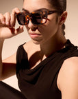 Eine Frau mit dunklem Haar trägt ein ärmelloses schwarzes Oberteil und eine polarisierte Sonnenbrille von Okkia. Sie blickt ernst nach vorne, während sie mit einer Hand die Sonnenbrille vor einem sanft beleuchteten neutralen Hintergrund hält.