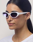 Eine Frau mit langen, nach hinten gebundenen dunklen Haaren trägt eine ovale weiße Okkia-Sonnenbrille mit UV 400 Schutz und ein weißes Hemd. Sie blickt leicht nach links vor einem einfarbigen, hellen Hintergrund, auf dem Sonnenbrillenbügel ist "Okkia" zu sehen.