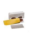 A pair of round, white-framed Okkia sunglasses with brown lenses sits next to a yellow case and gray cleaning cloth labeled “OKKIA.” Behind them is a beige Okkia box with a white cutout design, all displayed on a white background.