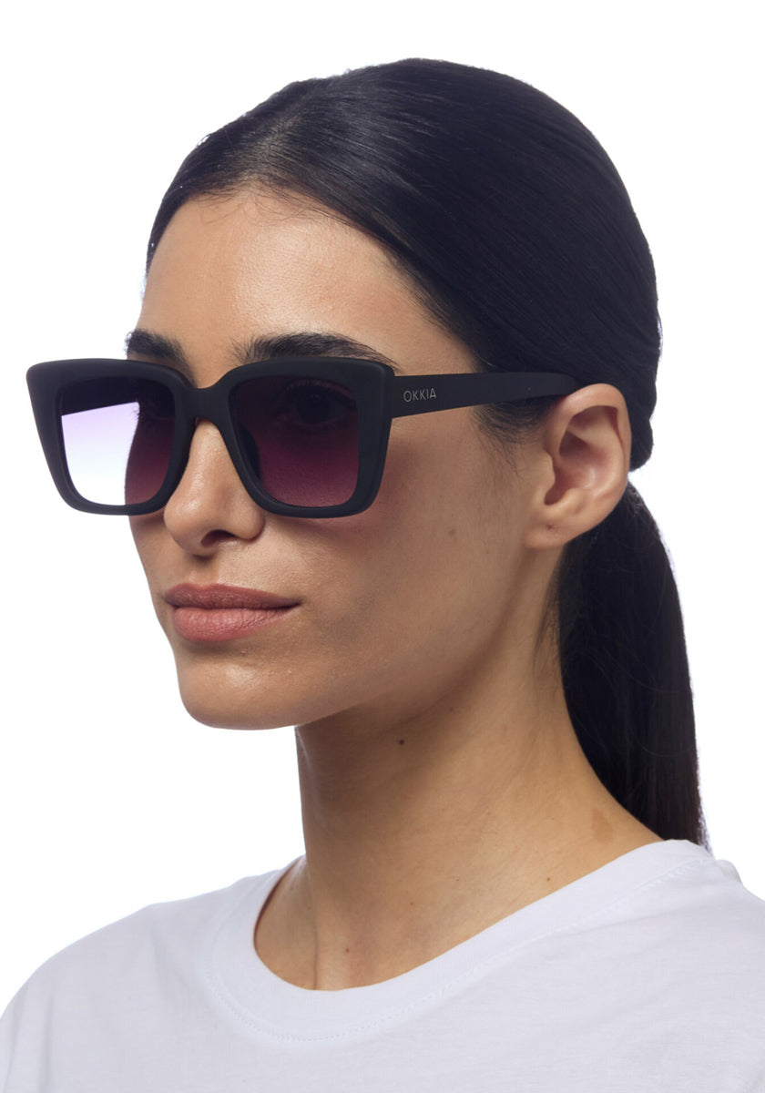 A woman with straight, dark hair pulled back wears large, square Okkia sunglasses with black frames and gradient lenses. She’s in a plain white top, facing slightly right against a white background. The Okkia logo appears on the temple.