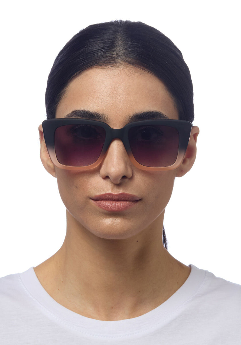 A person with dark, straight hair pulled back wears Okkia’s large square black sunglasses with gradient lenses. They have light skin, a neutral expression, and wear a white crew-neck shirt against a plain white background.