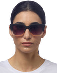 A person with dark, straight hair pulled back wears Okkia’s large square black sunglasses with gradient lenses. They have light skin, a neutral expression, and wear a white crew-neck shirt against a plain white background.