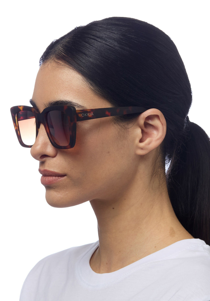 A woman with straight dark hair tied in a low ponytail wears Okkia oversized square tortoiseshell sunglasses with gradient brown lenses. She is shown in profile against a white background, dressed in a plain white T-shirt.