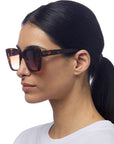 A woman with straight dark hair tied in a low ponytail wears Okkia oversized square tortoiseshell sunglasses with gradient brown lenses. She is shown in profile against a white background, dressed in a plain white T-shirt.