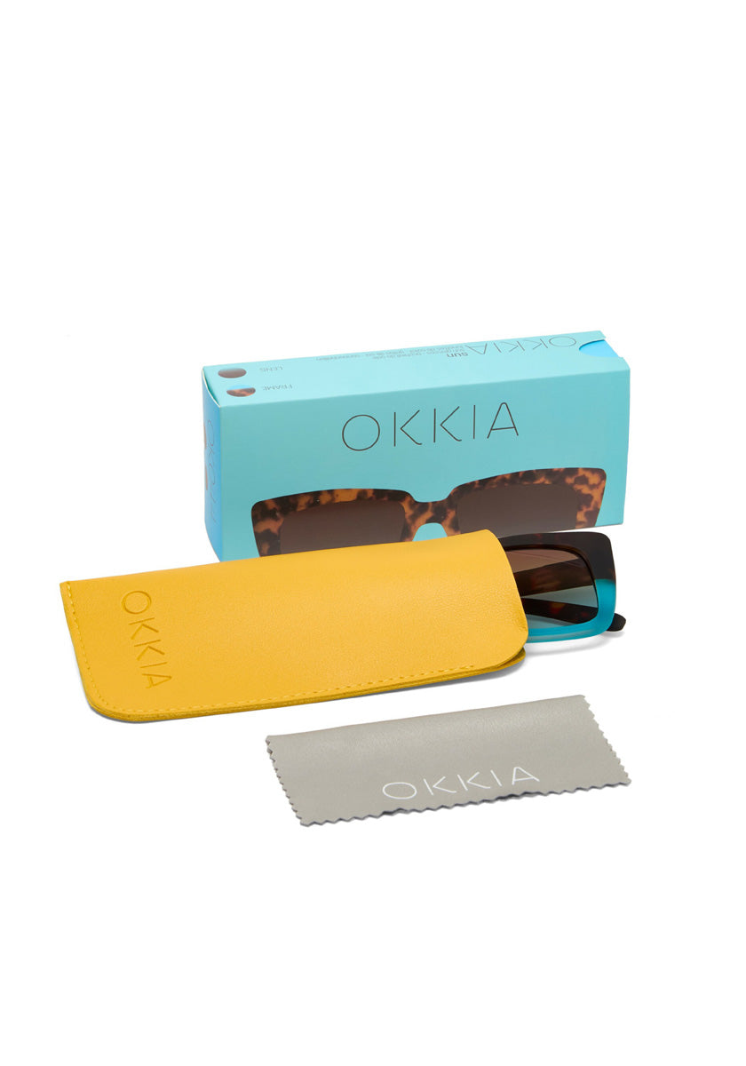 A pair of Okkia sunglasses with tortoiseshell frames sits partly in a bright yellow case. In front is a gray microfiber cloth with Okkia branding, and behind them is a blue Okkia box. All items are displayed on a white background.
