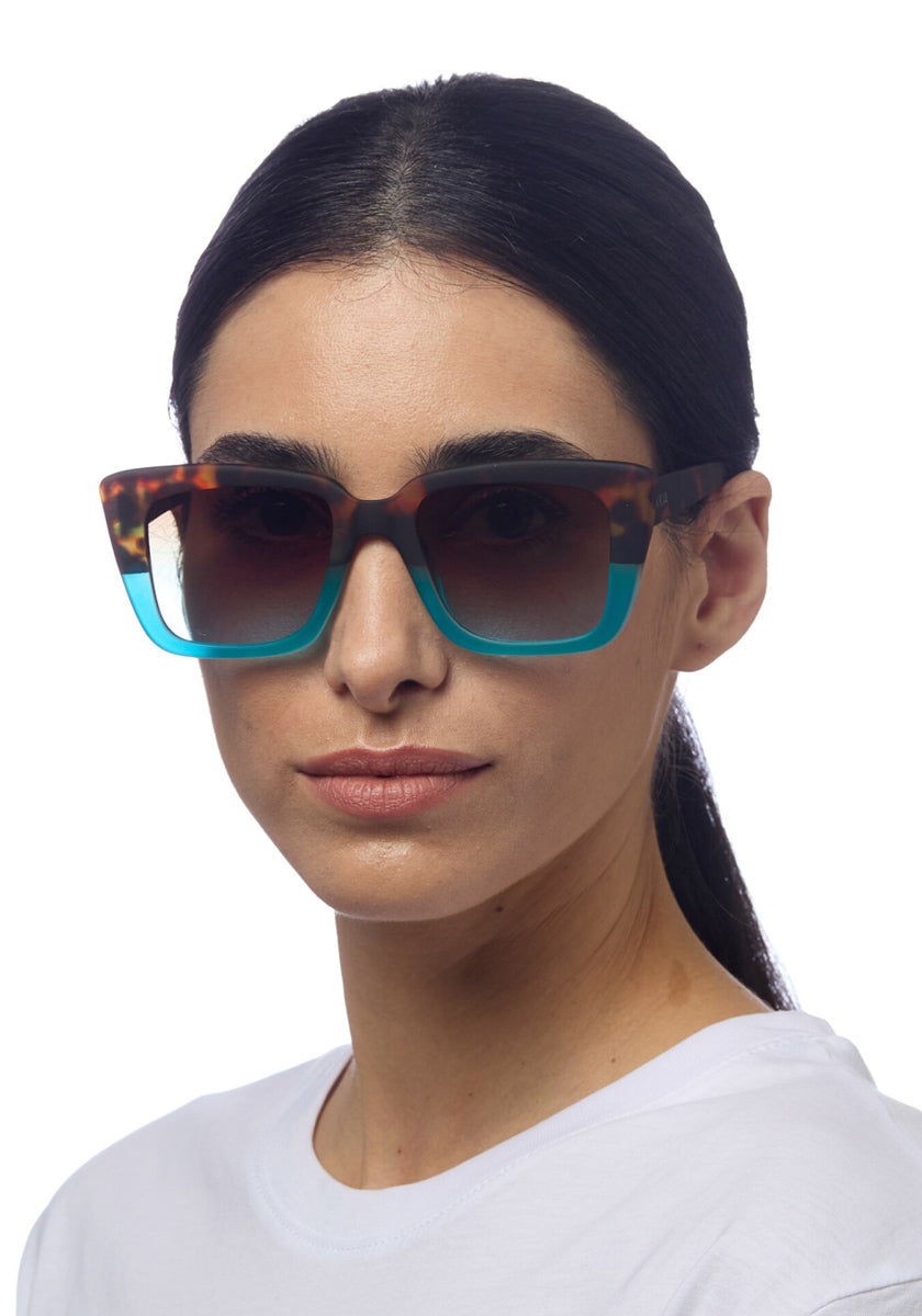 A woman with dark hair pulled back wears a white shirt and Okkia large square sunglasses featuring brown tortoiseshell frames that fade to teal blue at the bottom, facing slightly right against a white background.