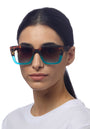 A woman with dark hair pulled back wears a white shirt and Okkia large square sunglasses featuring brown tortoiseshell frames that fade to teal blue at the bottom, facing slightly right against a white background.