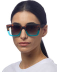 A woman with dark hair pulled back wears a white shirt and Okkia large square sunglasses featuring brown tortoiseshell frames that fade to teal blue at the bottom, facing slightly right against a white background.