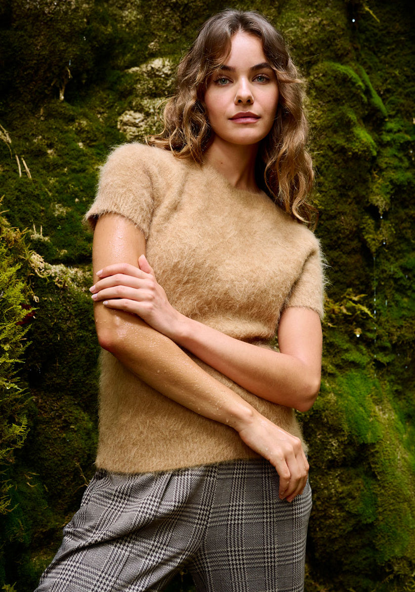 A woman stands in front of a lush, mossy wall. She has wavy brown hair and wears a short-sleeved, fuzzy camel-colored sweater and gray plaid pants. Her arms are crossed gently in front of her, and she looks calmly at the camera.