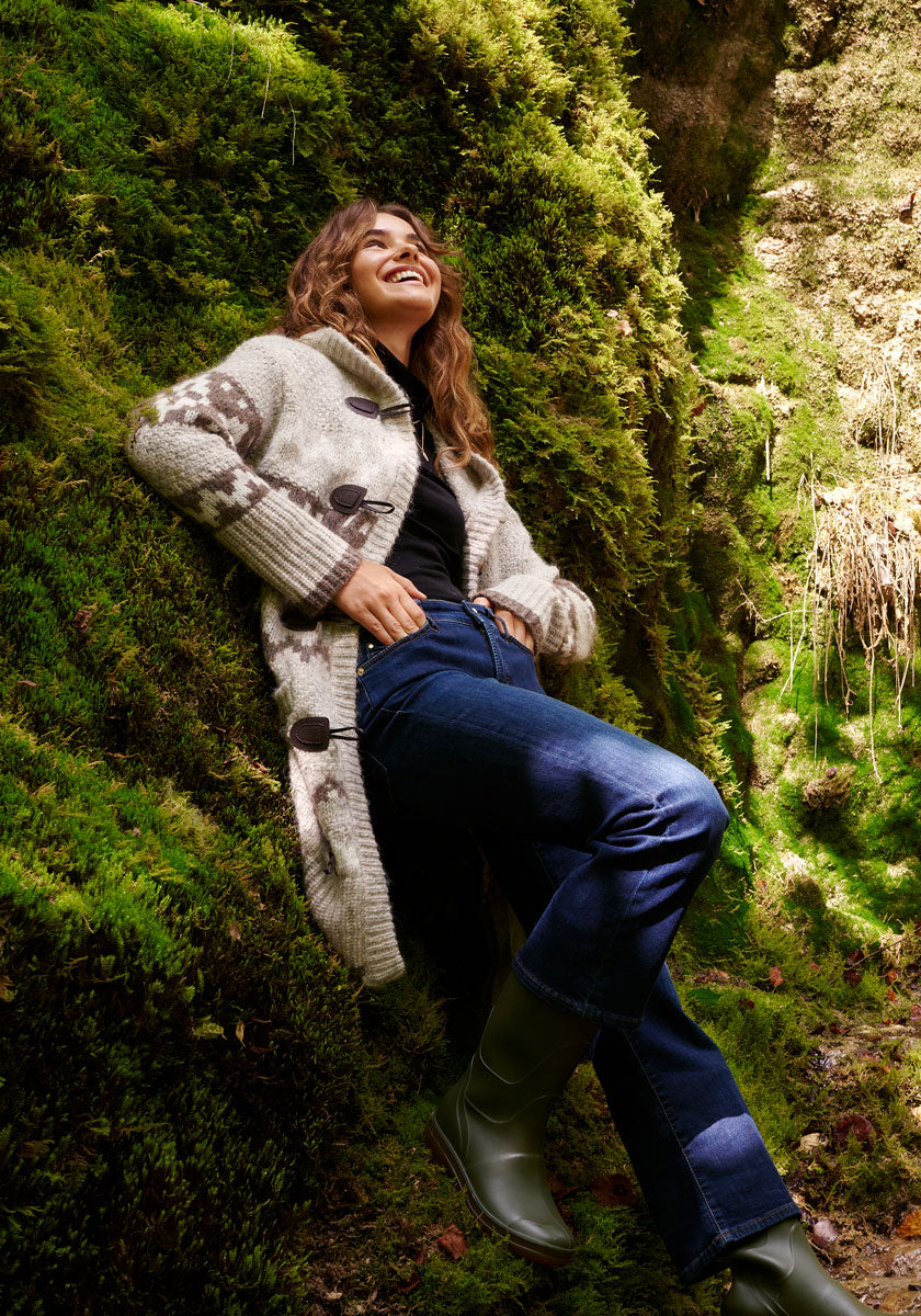 A woman with wavy hair leans against a moss-covered rock wall outdoors, smiling. She wears a patterned knit coat, dark jeans, black top, and green boots. Sunlight filters through, highlighting the lush green moss and her relaxed, joyful expression.