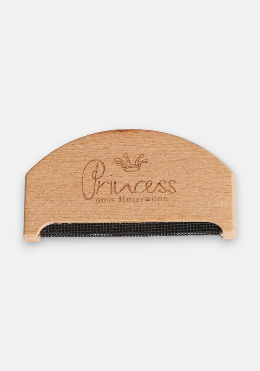 A semicircular Princess Goes Hollywood cashmere comb lint remover with a metal mesh flat edge. Princess goes Hollywood and a crown are engraved above the mesh. Perfect for removing fabric pilling, shown on a plain white background.