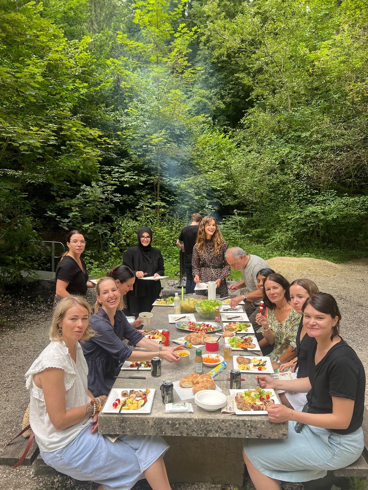 A group of people sit and stand around a picnic table outdoors in a lush, green forest. The table is filled with plates of food, drinks, and snacks. Some people are smiling at the camera, while others are serving food or chatting. Smoke rises in the background.