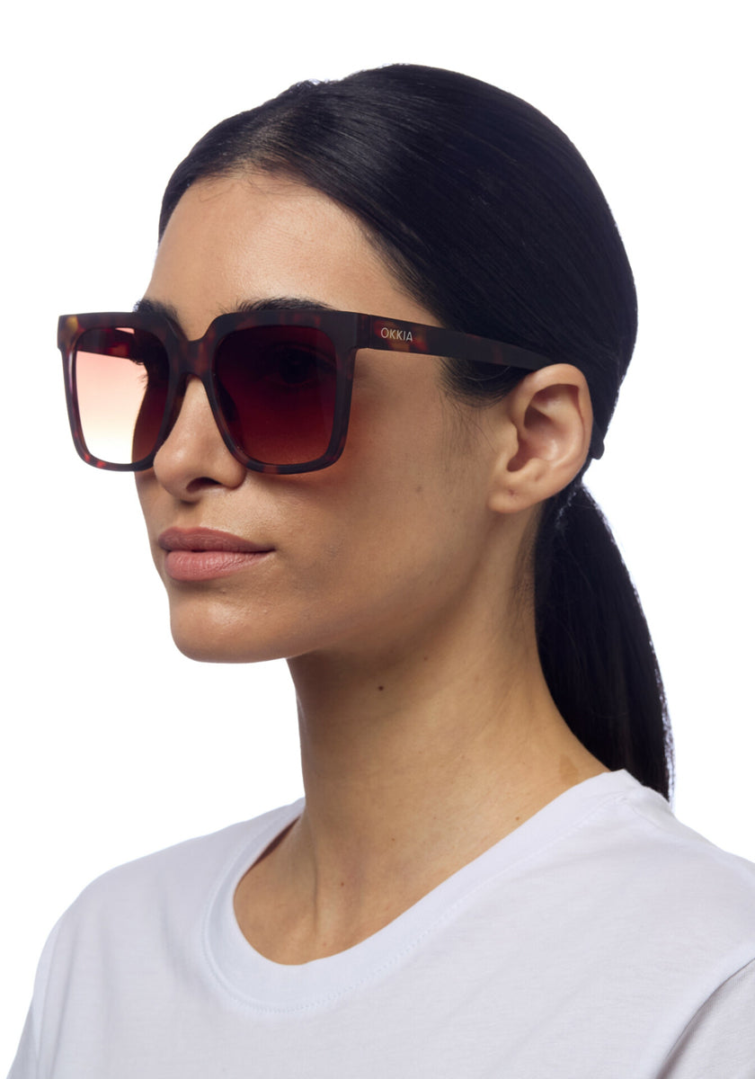 A woman with straight dark hair in a low ponytail wears oversized, square brown tortoiseshell sunglasses with gradient lenses and “Okkia” branding. She has a neutral expression and wears a plain white T-shirt against a white background.