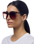 A woman with straight dark hair in a low ponytail wears oversized, square brown tortoiseshell sunglasses with gradient lenses and “Okkia” branding. She has a neutral expression and wears a plain white T-shirt against a white background.