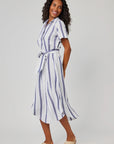 A woman with long curly hair smiles in a Bella Dahl Leinenmix Hemdkleid in Midi-Länge with vertical blue stripes and a matching Taillengürtel, paired with tan sandals, standing against a plain light gray background.