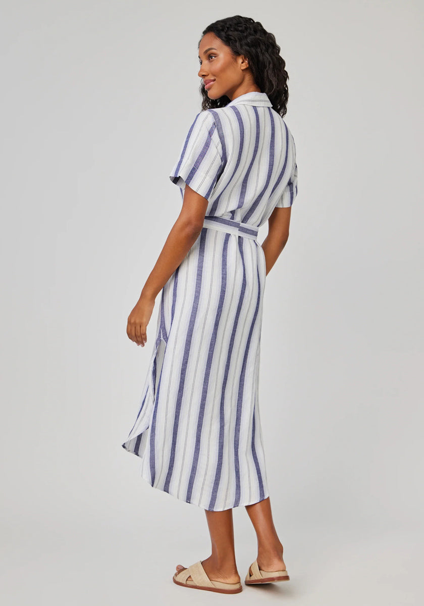 A woman with wavy dark hair stands against a light gray background, wearing a blue-and-white striped Bella Dahl midi-length linen-blend shirtdress with a waist belt and light sandals.
