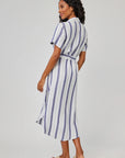 A woman with wavy dark hair stands against a light gray background, wearing a blue-and-white striped Bella Dahl midi-length linen-blend shirtdress with a waist belt and light sandals.