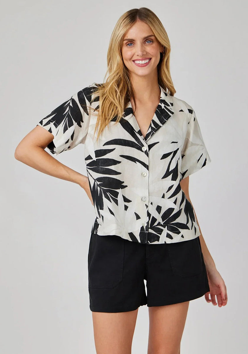 A smiling woman with long blonde hair wears a Bella Dahl short-sleeve, button-up palm print blouse on white fabric with black shorts, standing against a plain light background with one hand on her hip.