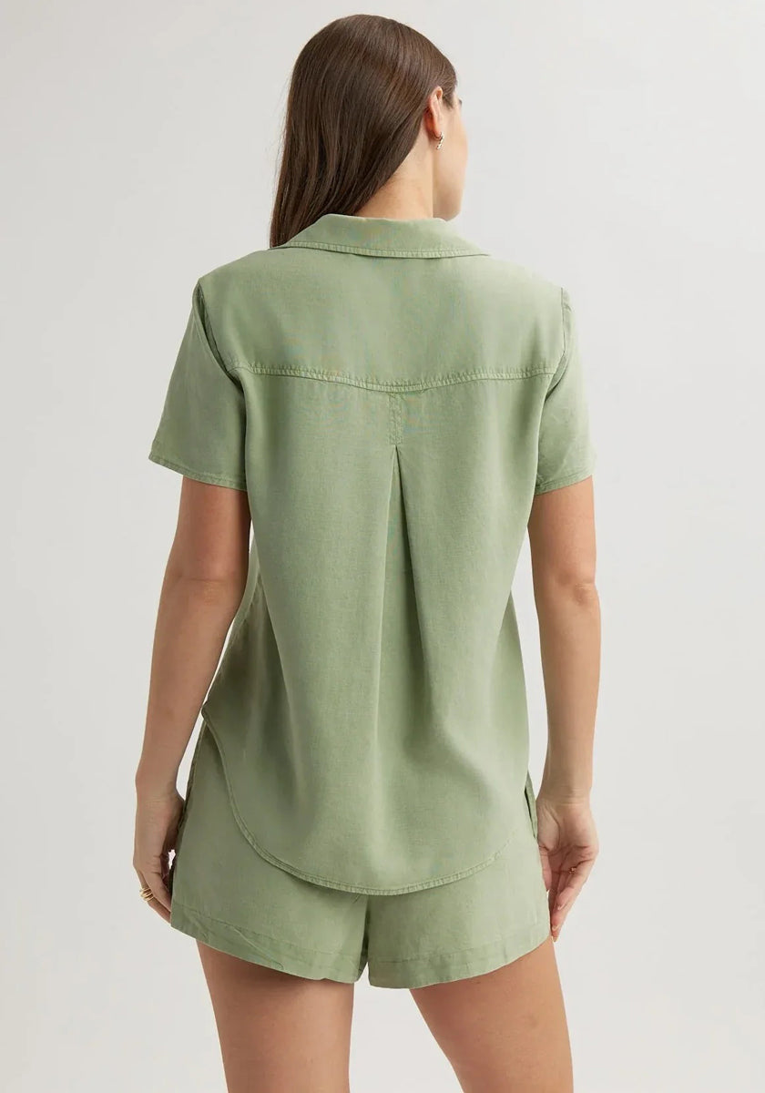 A woman with long brown hair faces away, wearing a sage green Bella Dahl Lyocell short-sleeve shirt with Hemdkragen and matching loose shorts. The shirt has a back yoke with pleat and a curved hem. The background is plain and light-colored.