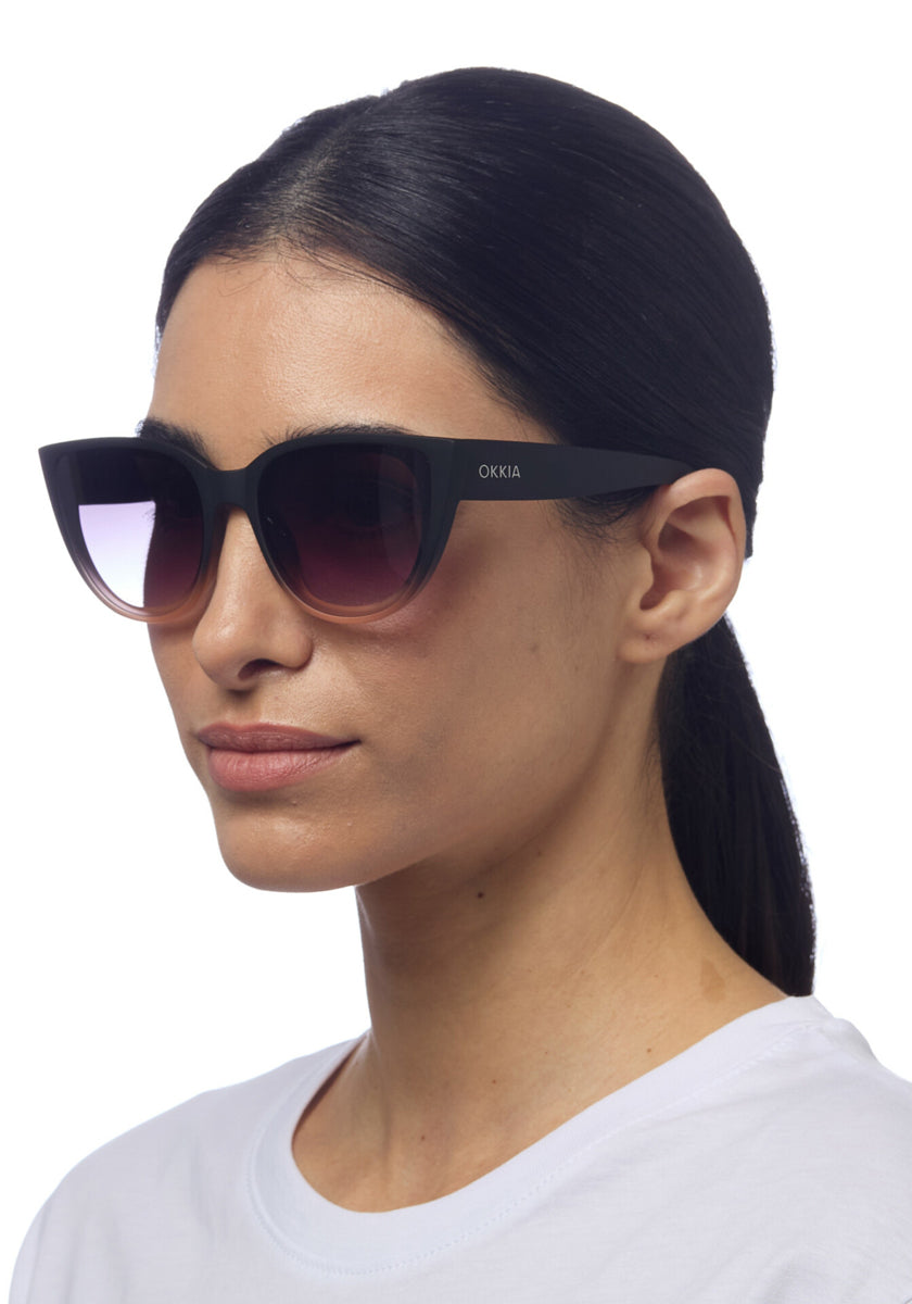 A woman with straight dark hair tied back wears oversized cat-eye sunglasses by Okkia with gradient lenses and UV protection. She faces slightly left in a plain white top against a white background, her expression neutral.