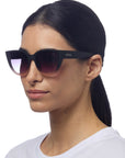 A woman with straight dark hair tied back wears oversized cat-eye sunglasses by Okkia with gradient lenses and UV protection. She faces slightly left in a plain white top against a white background, her expression neutral.