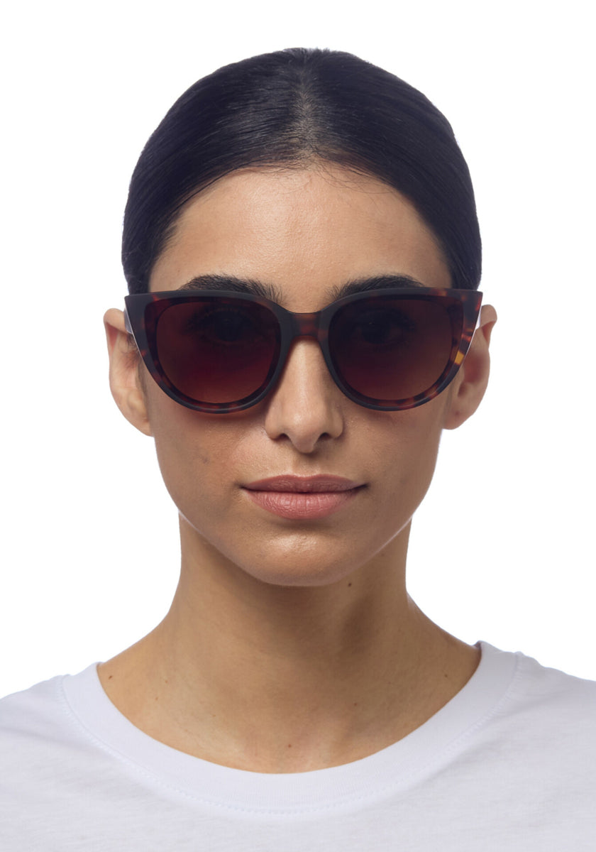 A woman with straight dark hair wears Okkia large, round sunglasses featuring Soft-Touch-Oberfläche and UV-Schutz, paired with a white crew-neck shirt. She faces forward against a plain white background, her expression neutral.
