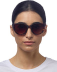 A woman with straight dark hair wears Okkia large, round sunglasses featuring Soft-Touch-Oberfläche and UV-Schutz, paired with a white crew-neck shirt. She faces forward against a plain white background, her expression neutral.