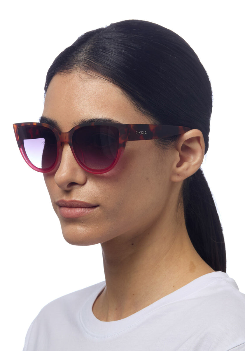 A woman with straight dark hair in a low ponytail wears oversized Okkia cat-eye sunglasses with tortoiseshell frames, soft-touch finish, and gradient pink lenses. She has a neutral expression, dressed in white against a white background.