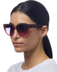 A woman with straight dark hair in a low ponytail wears oversized Okkia cat-eye sunglasses with tortoiseshell frames, soft-touch finish, and gradient pink lenses. She has a neutral expression, dressed in white against a white background.