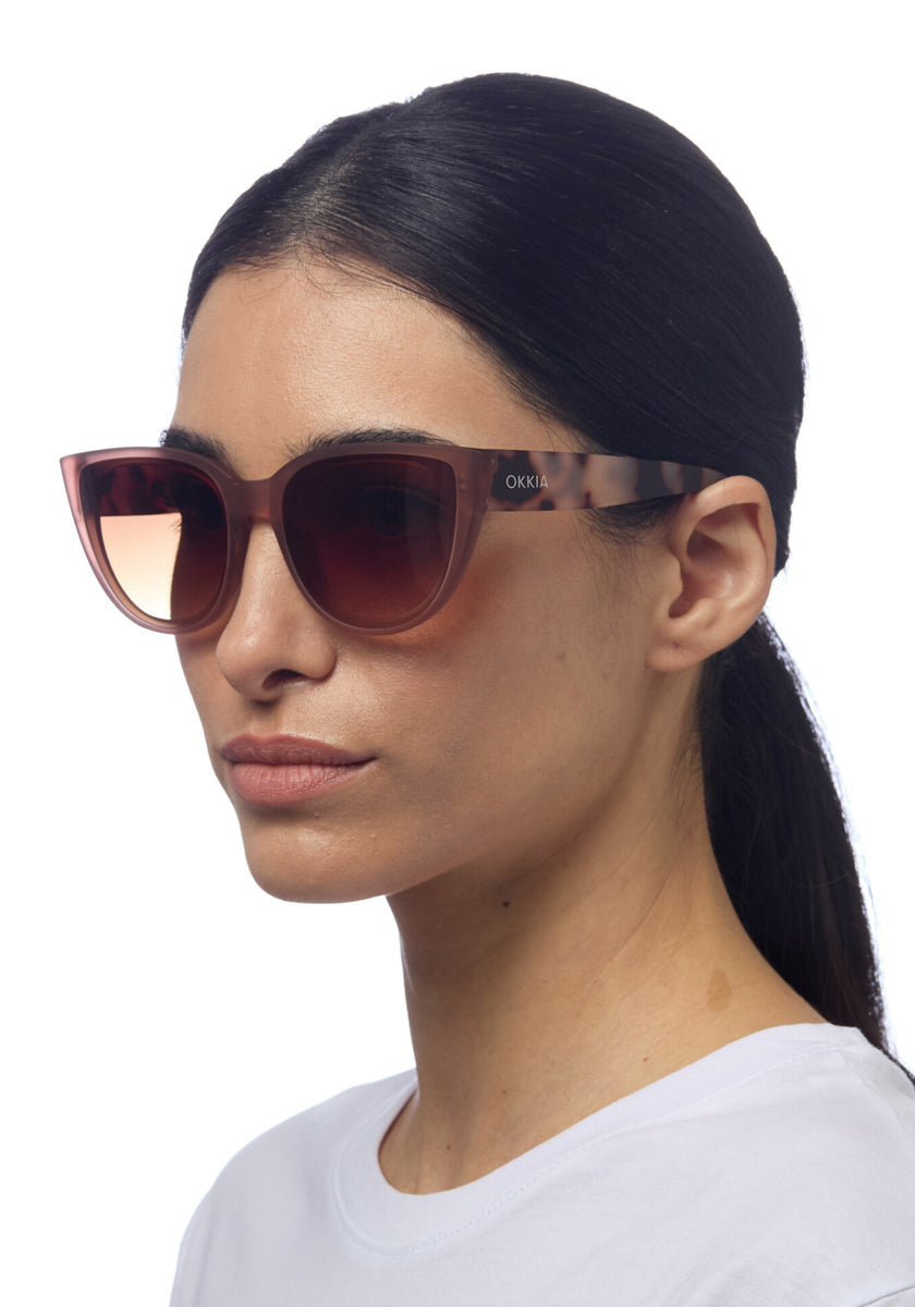 A woman with straight dark hair pulled back wears oversized Okkia cat-eye sunglasses with pink-gradient lenses and thick soft-touch frames. Okkia is visible on the side. She is in profile, facing left, in a white top against a white background.