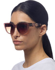 A woman with straight dark hair pulled back wears oversized Okkia cat-eye sunglasses with pink-gradient lenses and thick soft-touch frames. Okkia is visible on the side. She is in profile, facing left, in a white top against a white background.