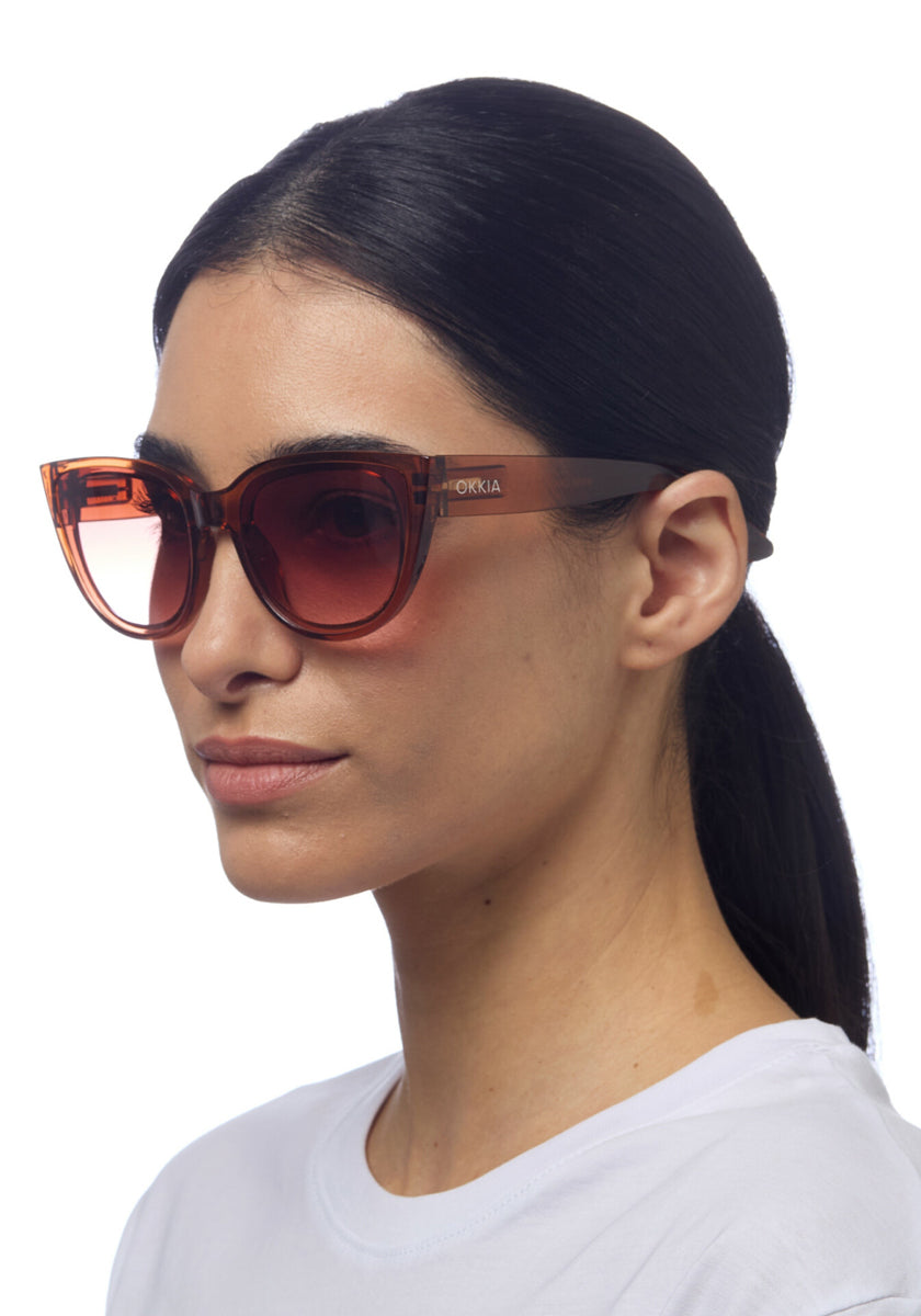 A woman with straight dark hair tied back wears oversized, brown-tinted Cat-Eye-Sonnenbrille by Okkia with thick semi-transparent frames and UV-Schutz, looking slightly to the side against a plain white background.
