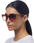 A woman with straight dark hair tied back wears oversized, brown-tinted Cat-Eye-Sonnenbrille by Okkia with thick semi-transparent frames and UV-Schutz, looking slightly to the side against a plain white background.