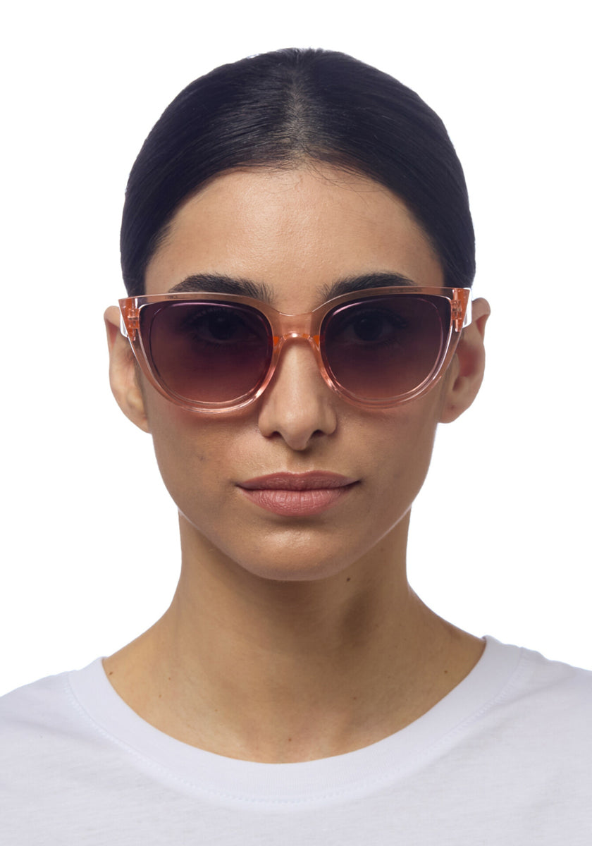 A woman with dark hair parted in the center wears translucent pink Okkia cat-eye sunglasses and a white crew-neck shirt. She faces forward against a plain white background, showing a neutral expression.