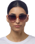 A woman with dark hair parted in the center wears translucent pink Okkia cat-eye sunglasses and a white crew-neck shirt. She faces forward against a plain white background, showing a neutral expression.