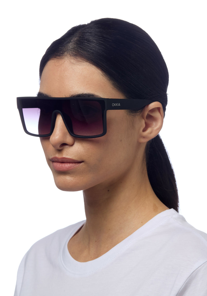 A woman with straight dark hair tied back wears a white shirt and oversized Okkia sunglasses featuring rectangular black frames, gradient lenses, and UV 400 protection. The Okkia logo is visible as she faces slightly left against a white background.