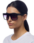 A woman with straight dark hair tied back wears a white shirt and oversized Okkia sunglasses featuring rectangular black frames, gradient lenses, and UV 400 protection. The Okkia logo is visible as she faces slightly left against a white background.