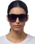 A woman with straight dark hair pulled back wears oversized, rectangular Okkia sunglasses with UV 400 protection and a plain white shirt. She faces forward against a white background, showing a neutral facial expression.