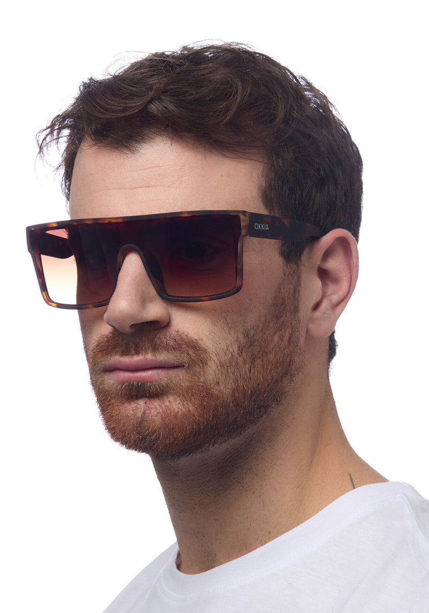 A man with short brown hair and a trimmed beard wears Okkia Sonnenbrille im Oversized-Stil mit UV 400-Schutz und Verlaufsgläsern. He has a neutral expression in a white crew-neck shirt, posing against a plain white background.