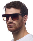 A man with short brown hair and a trimmed beard wears Okkia Sonnenbrille im Oversized-Stil mit UV 400-Schutz und Verlaufsgläsern. He has a neutral expression in a white crew-neck shirt, posing against a plain white background.