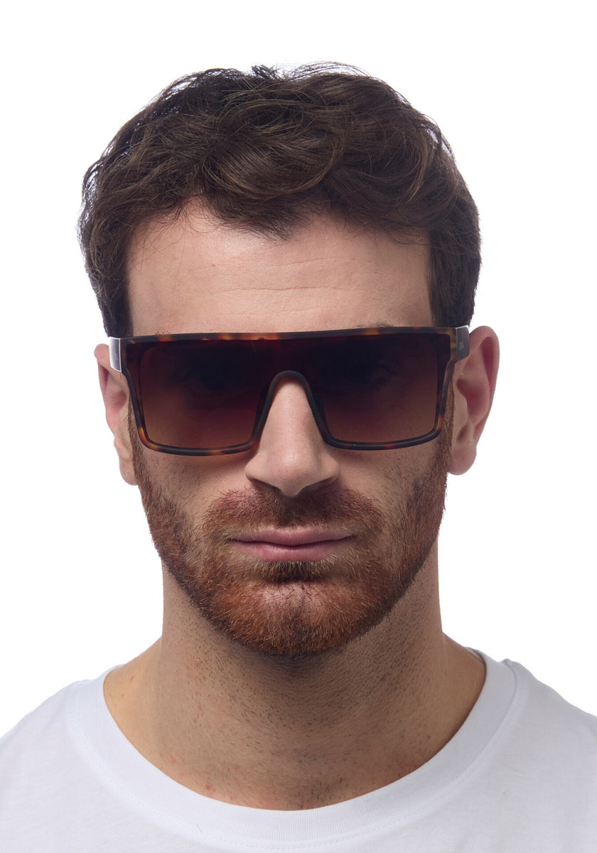 A man with short brown hair and a full reddish-brown beard wears oversized Okkia Sonnenbrille with large, square brown frames and UV 400 protection. He faces forward in a plain white T-shirt against a white background, his expression neutral.