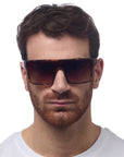 A man with short brown hair and a full reddish-brown beard wears oversized Okkia Sonnenbrille with large, square brown frames and UV 400 protection. He faces forward in a plain white T-shirt against a white background, his expression neutral.