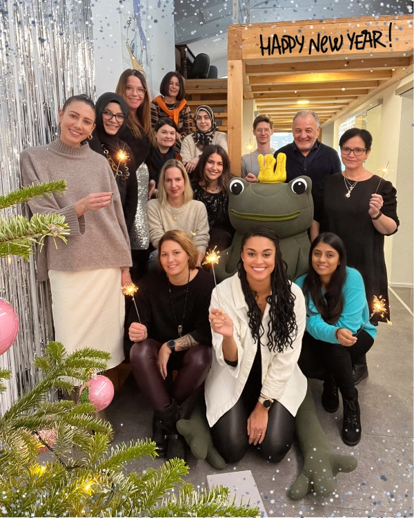 A group of people, smiling and holding sparklers, pose indoors beside a large frog mascot wearing a crown. A decorated Christmas tree is on the left, and a “Happy New Year!” sign hangs above. Silver tinsel decorates the background. Snowflake graphics overlay the image.
