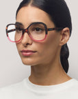 Eine Frau mit glattem, dunklem Haar trägt eine große, runde Okkia Lesebrille mit einem schwarz-rosa Schmetterlingsrahmen. Sie hat einen neutralen Ausdruck, ist weiß gekleidet und steht vor einem hellgrauen Hintergrund.
