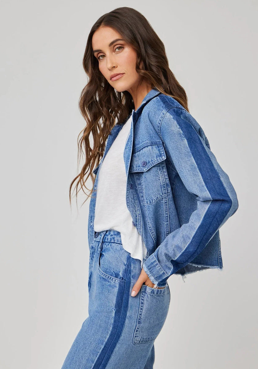 A woman with long, wavy brown hair poses sideways in a Bella Dahl denimoptik jacket and high-waist hose with blue side stripes, paired with a white t-shirt. Her left hand is in her pocket. The plain light gray background highlights the lyocell blend outfit.