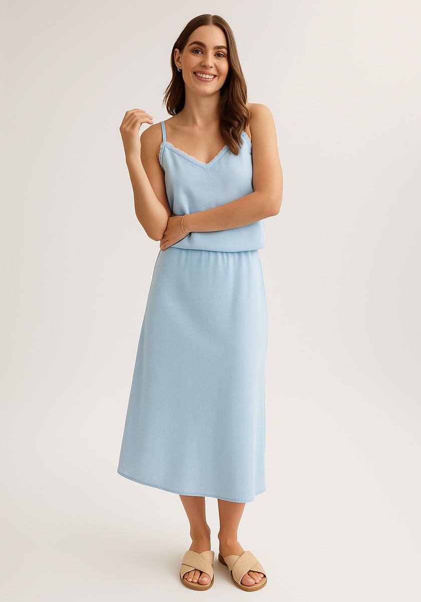 Against a plain background, a woman smiles confidently in a light blue Bella Dahl Lyocell midi dress with thin straps and an A-line skirt, paired with beige slide sandals. Her loose, wavy brown hair enhances her relaxed pose.