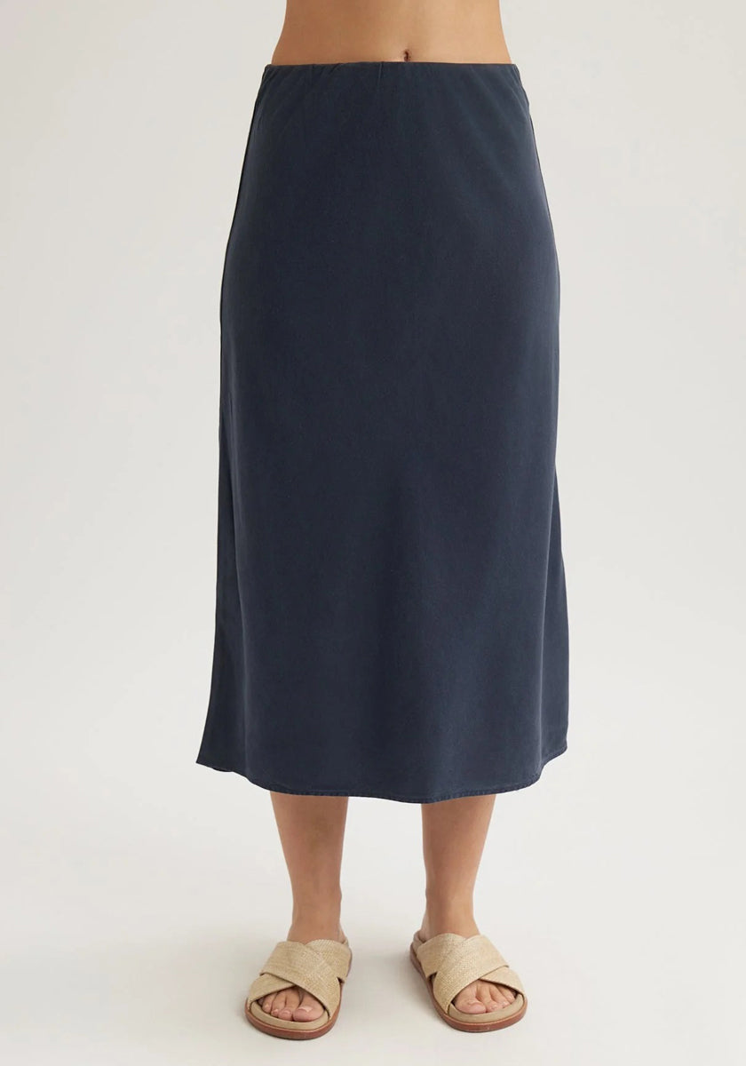 A persons lower torso and legs are shown against a plain light background, wearing a Bella Dahl navy blue midi flared skirt and beige open-toed sandals with crisscross straps.